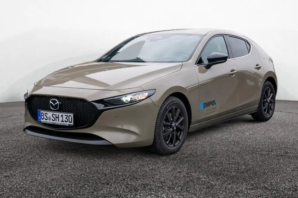 Seitlich-frontale Sicht auf einen Mazda 3 in der Farbe platinum-quartz, der auf dem Hof des Autohauses Hempel in Braunschweig steht.